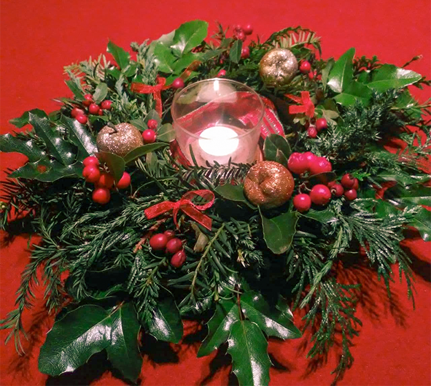 Ein winterlicher Kranz mit einer Kerze in der Mitte. Der Kranz ist aus Thuja, Ilex und Tanne gebunden. Geschmückt ist er mit roten Beeren, goldenen Dekoäpfeln und roten Schleifchen.