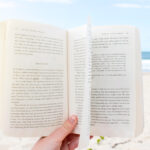 Ein aufgeschlagenes Buch wird in die Kamera gehalten, im Hintergrund ein Strand und ein Meer