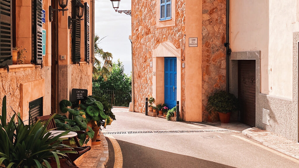 Eine sonnenbeschienene Straße auf Mallorca mit orangenen Häusern, die blaue Türen und Fensterrahmen haben.