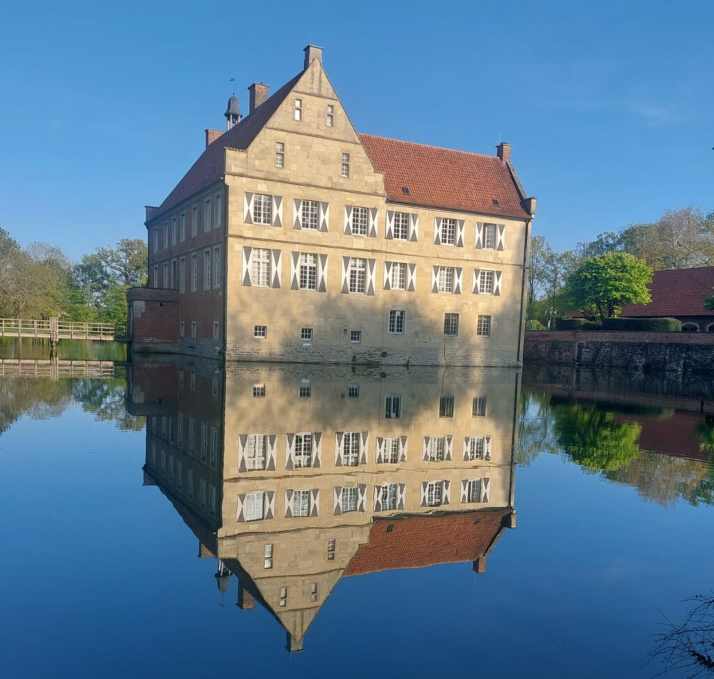 Burg Hülshoff spiegelt sich in der Gräfte. Rechts daneben steht ein Teil der Wirtschaftshäuser.