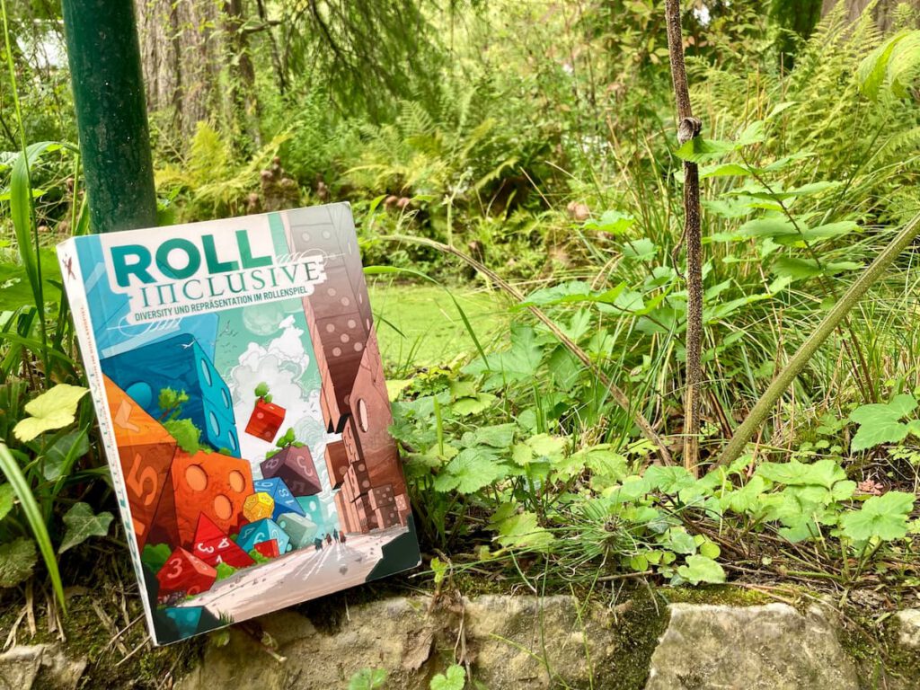 Das Buch "Roll Inclusive" lehnt an einem Baum auf einer moosbbewachsenen Mauer. Im Hintergrund ein grüner Wald.