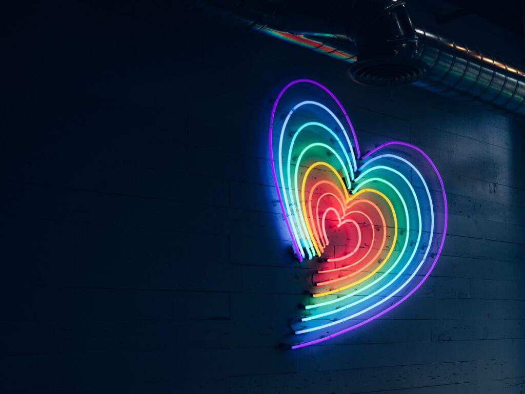 Eine Reihe von schmalen leichtenden Neonröhren auf einer Betonwand ergeben ein Herz in Regenbogenfarben.