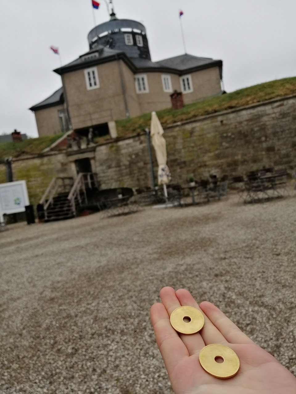 Eine Hand hält zwei goldene Wertmarken. Im Hintergrund ist die Festung Wilhelmstein zu sehen.